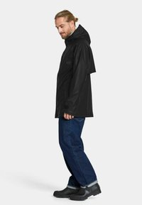 Svart vattentät regnjacka med huva, med rak skärning och justerbara manschetter, i kombination med blå jeans och svarta skor.