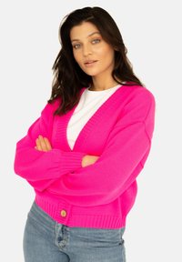 Fuksjowy, oversize kardigan z prążkowanymi mankietami i dołem, z głębokim dekoltem w kształcie litery V oraz jednym zapięciem na drewniany guzik, noszony na białej bluzce.