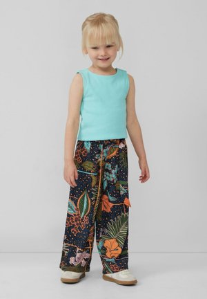 Jong meisje met blond haar in een mouwloze turquoise top, brede bloemenprintbroek en witte schoenen, staand tegen een effen achtergrond.