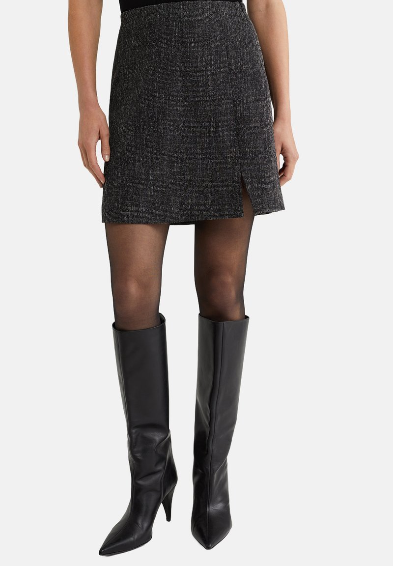 Jupe mini noire avec un motif texturé et une fente sur le côté, associée à des bottes en cuir noires montantes à bout pointu et à talon aiguille.