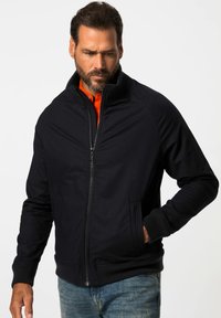 Svart zip-up jacka med hög krage, ribbade cuffs och sidofickor. Strukturerad tyg, i kombination med en ljusorange skjorta under.