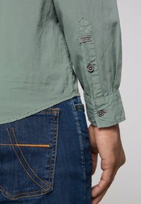Lichtgroene blouse met lange mouwen en knoopsluitingen op de manchetten, gecombineerd met blauwe spijkerbroeken met oranje stiksels op de zakken.
