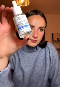 Kvinne i grå genser holder en flaske med Kiehl’s hudpleieserum nær kameraet under varmt innendørs lys.