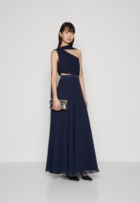 Robe de soirée bleu marine avec décolleté asymétrique, tissu drapé et jupe fluide. Assortie à une pochette métallique et des talons noirs.