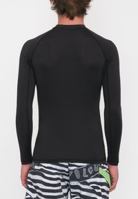 Czarna koszulka z długim rękawem typu rash guard, wykonana z elastycznego materiału, dopasowany krój, z płaskimi szwami. Noszona na pasiastych szortach kąpielowych.