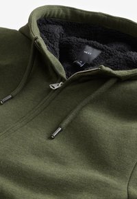 Grüner Reißverschluss-Hoodie mit einem strukturierten schwarzen Fleece-Innenfutter, ausgestattet mit einem Metallreißverschluss und Kordeln, aus einem weichen Stoff gefertigt.