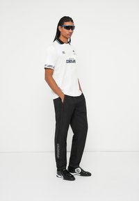 Witte T-shirt met korte mouwen en een zwarte kraag, voorzien van het "TRUE COLOURS" logo, gecombineerd met zwarte broek met witte zijnaad en logo-accenten.
