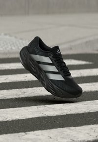 Μαύρο αθλητικό παπούτσι Adidas με λευκές ρίγες στον αέρα πάνω από διάβαση πεζών, με μικρά σωματίδια βρωμιάς γύρω από την σόλα.
