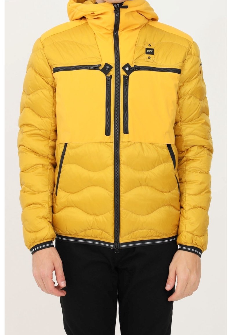 Veste matelassée jaune avec une texture capitonnée, dotée d'une fermeture éclair à l'avant, de garnitures noires et d'une capuche. Deux poches poitrine et des poches latérales.