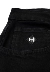 Sort denimstof med synlige sømme og en baglomme med et diamantformet broderet logo i hvid.