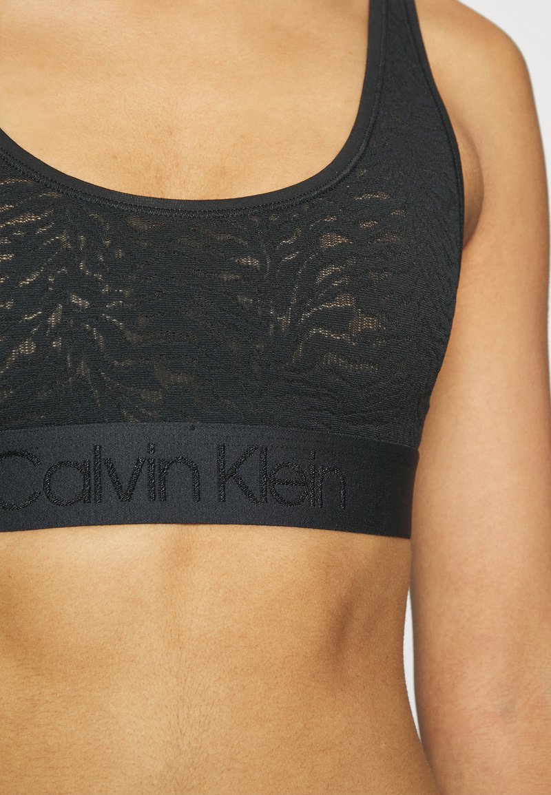 Černý krajkový bralet s texturovaným vzorem a širokým elastickým páskem s kontrastním výšivkovým logem „Calvin Klein“. 