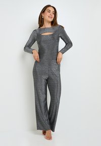 Combinaison métallique grise avec manches longues, présentant une découpe à l'avant, des jambes larges et des poches latérales. Tissu lisse et texturé avec un effet scintillant.