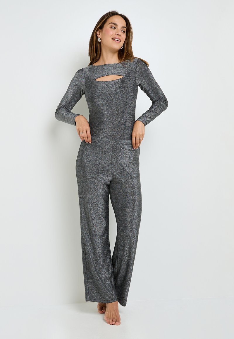 Combinaison métallique grise avec manches longues, présentant une découpe à l'avant, des jambes larges et des poches latérales. Tissu lisse et texturé avec un effet scintillant.