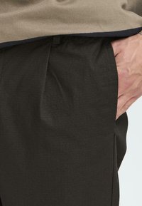 Una vista en primer plano de una mano de una persona descansando en el bolsillo de unos pantalones oscuros y texturizados, con una parte de una camiseta beige visible encima.