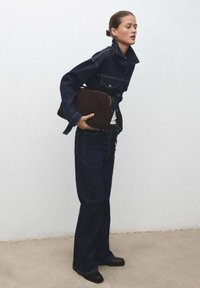 Tamno plavi traper jakna s zakrpanim džepovima, uparen s odgovarajućim flared farmerkama. Drži smeđu kožnu torbu, postavljen na jednoličnu pozadinu.