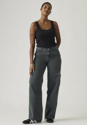 Kobieta stojąca, ubrana w czarny top na ramiączkach, ciemnoszare szerokie spodnie typu cargo, czarne trampki, z tatuażem na lewej górnej ramieniu i srebrną biżuterią.