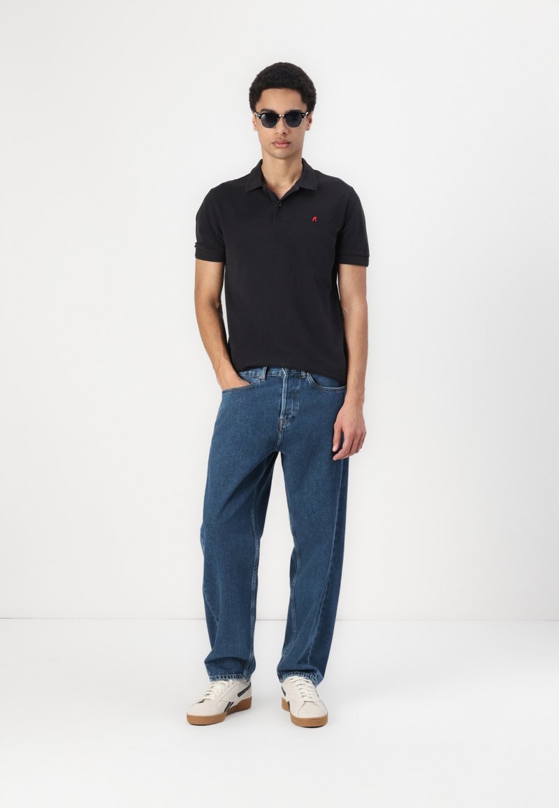 Polo negro de manga corta con un logo rojo, combinado con unos vaqueros de mezclilla azul holgados y zapatos blancos con suela de color marrón claro. Modelo usando gafas de sol.