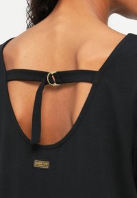 Μαύρο τοπ με ανοιχτή πλάτη, λουράκι και κυκλική χρυσή λεπτομέρεια. Το ύφασμα φαίνεται μαλακό με λεία υφή. Ο ετικέτα Barbour είναι ορατή.