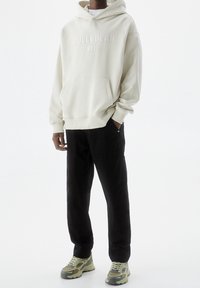 Persona che indossa un hoodie crema con la scritta "PULL&BEAR", pantaloni neri e sneakers beige-verdi, in piedi contro uno sfondo bianco uniforme.