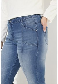 Tummansiniset farkut, joissa on hieman venyvyyttä, keski- korkea vyötärö ja takatasku, jossa on pieni metallinen napin yksityiskohta. Sujuva pinta, istuva siluetti.