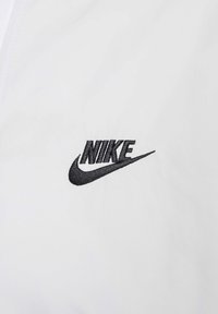 Biały materiał o gładkiej fakturze z czarnym haftowanym logo Nike. Logo zawiera stylizowane litery oraz znak „swoosh”.