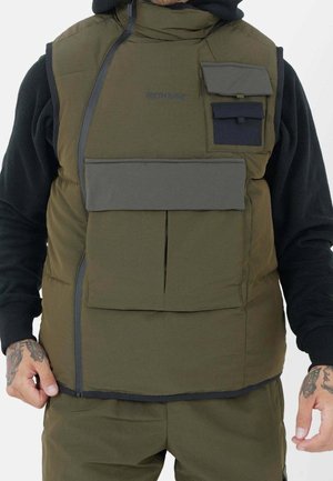 Homme portant un gilet matelassé vert olive avec une fermeture éclair chevauchante et plusieurs poches à rabat sur des manches longues noires, montrant des mains tatouées.