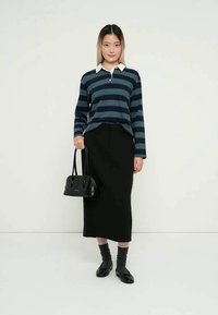 Jovem mulher vestindo uma camisa de mangas longas azul e verde com listras, saia midi preta, sapatos pretos, meias cinzas, segurando uma bolsa preta.