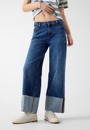 Szerokie niebieskie dżinsy z postrzępionym, jasnoniebieskim mankietem. Wykonane z mocnej tkaniny, z tradycyjnymi kieszeniami i zapięciem na guzik.