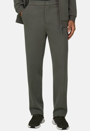 Homme portant un pantalon droit vert foncé avec une poche zippée sur le côté et des chaussures de sport noires.
