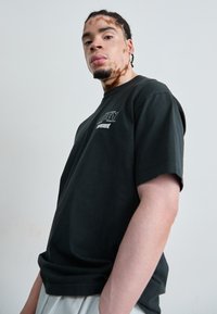 T-shirt noir à manches courtes avec un col rond, arborant un logo en texte blanc sur la poitrine. Le tissu semble doux avec une coupe décontractée.