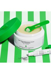 Weißes Glas mit einem grünen Deckel, das einen cremigen Butterreiniger enthält. Ein grüner Löffel ist enthalten. Der Hintergrund besteht aus grünen und weißen Streifen.