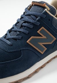 Mörkblå sneakers i mockamaterial, med en framträdande logotyp i brunt läder, texturerad tygtunga och ljus sula med bra grepp.