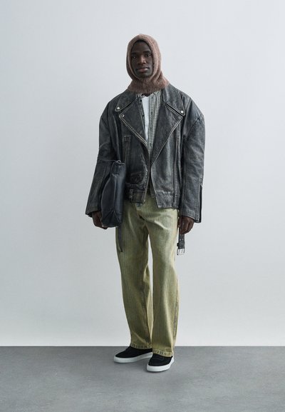 Muž v hnedej pletenej kapucni, nadrozmernej čiernej koženej bunde, károvanej košeli, voľných žltých džínsoch, čiernych teniskách, držiaci čiernu koženú tašku, stojaci na sivej podlahe.