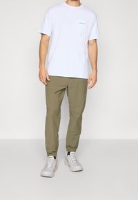 Man i en vit Patagonia t-shirt, olivgröna joggingsbyxor och vita Columbia sneakers, som står mot en enkel bakgrund.