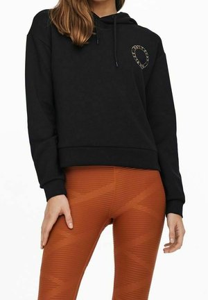 Kvinde iført en sort hættetrøje med et lille grafik på brystet og strukturerede brændt orange leggings.