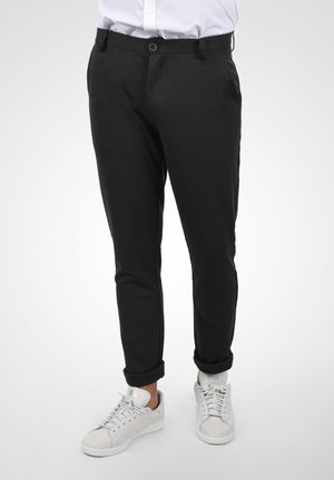Sorte tapered bukser lavet af glat stof, med knaplukning, bæltestropper og opsmøg ved anklerne, parret med hvide sneakers.