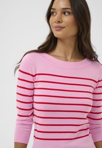 Femme portant un pull rose à rayures rouges horizontales, regardant sur le côté, avec de longs cheveux bruns et un collier argenté.