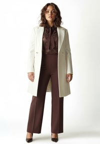 Weißer, maßgeschneiderter Mantel mit zwei Knöpfen, übereinander getragen über einer braunen Bluse mit hohem Kragen und passenden Hosen, vervollständigt durch schwarze Lacklederschuhe.