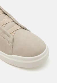 Beige Wildleder-Sneaker mit runder Zehenpartie, elastischen Schnürsenkeln und einer weißen Gummisohle. Besitzt dezente Nähte und eine glatte Textur.