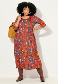 Robe maxi en orange vif avec des motifs en plumes multicolores. Présente des manches trois-quarts et une coupe décontractée, accompagnée de bottes marron.