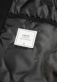 Black quilted fabric with a smooth texture, featuring a white label for "next TAILORING," size M, stitched into the interior.