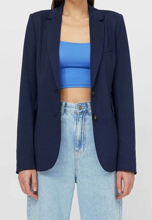 Persoon die een marineblazer draagt over een blauw crop top, gecombineerd met een lichtblauwe spijkerbroek met hoge taille, staand tegen een effen achtergrond.