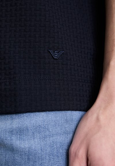 Top de tecido encaixado azul marinho com um logotipo bordado na bainha. A peça é combinada com jeans lilás, destacando contrastes de textura.
