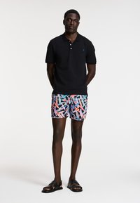 Čierna polo košeľa, farebné plavky s vzorom, čierne sandále. Má krátke rukávy, klasický golier a živý geometrický dizajn.