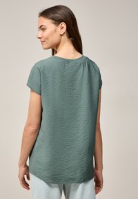 Top verde a maniche corte con tessuto texturizzato, scollatura rotonda e orlo posteriore leggermente più lungo. Vestibilità casual e comoda.