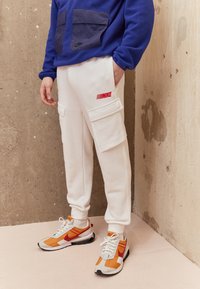 Vita cargobyxor i sweatpant-stil med två sidofickor och röd "NIKE"-logotyp, kombinerat med orange och gråa träningsskor samt en blå fleece-tröja.