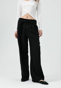 Zwarte broek met wijde pijpen en een taille met riem, gemaakt van gladde stof. Gecombineerd met een wit geribd shirt met lange mouwen en een overslagontwerp.