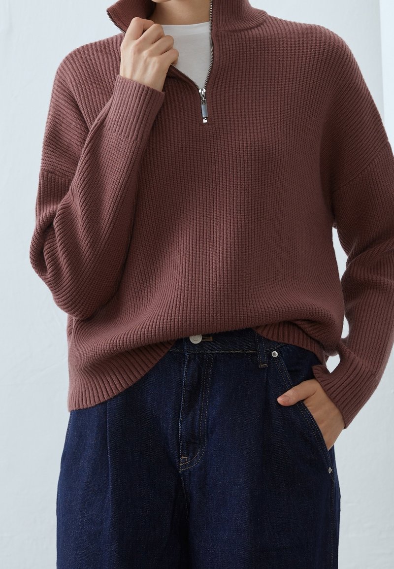 Personne portant un pull côtelé mauve à fermeture éclair quart de longueur sur une chemise blanche, avec un jean en denim bleu foncé, une main touchant le col et l'autre dans la poche.