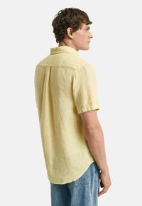 Kurzärmliges, hellgelbes Leinenhemd mit Kragen, Rückenpasse und abgerundetem Saum. Kombiniert mit lockeren Denim-Jeans.