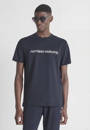 Hombre vistiendo una camiseta Antony Morato azul oscuro y pantalones a juego, con gafas de sol negras, posando frente a un fondo claro y liso.